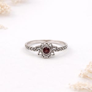 May include: A silver ring with a floral design and a round, deep red gemstone at the centre. The band features a textured pattern. The ring is set against a white background, highlighting its intricate details.