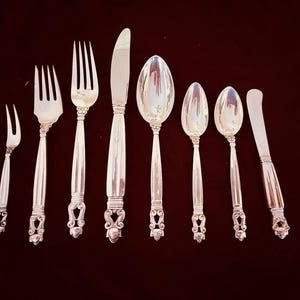 Op de afbeelding: Een verzameling zilverkleurig bestek, waaronder vorken, messen en lepels, gerangschikt op een donkere ondergrond. De gebruiksvoorwerpen hebben sierlijke handvatten met decoratieve details. De set bevat een verscheidenheid aan serveerstukken, geschikt voor een formele eetsituatie.