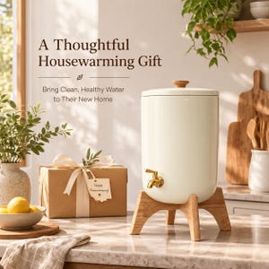 May include: A cream-coloured ceramic water dispenser with a gold-toned spigot and a wooden lid, resting on a light wood stand. Next to it is a brown gift box with a white ribbon and a tag reading "Happy Housewarming!" with text above.