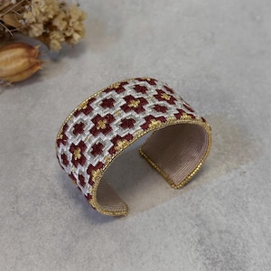 Puede incluir: Pulsera ancha con un patrón floral en burdeos, blanco y dorado. La pulsera tiene un ribete dorado y un interior beige. La pulsera mide aproximadamente 5 cm de ancho.