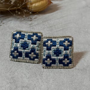 Puede incluir: Un par de pendientes cuadrados con un patrón geométrico. Los pendientes presentan un diseño en tonos azules y blancos, con un borde de cuentas plateadas. Un accesorio único.