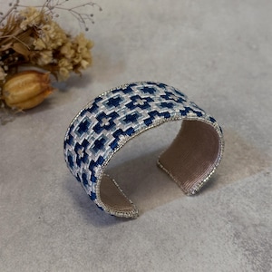 Puede incluir: Un brazalete ancho con un patrón geométrico en tonos azules y blancos. El brazalete tiene un borde plateado y un interior beige. El diseño es bordado y el brazalete parece hecho a mano.