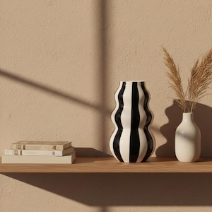 May include: A decorative shelf displays a black and white striped vase, a small white vase with dried plants, and a stack of books. The vases are on a wooden shelf against a neutral-toned wall.