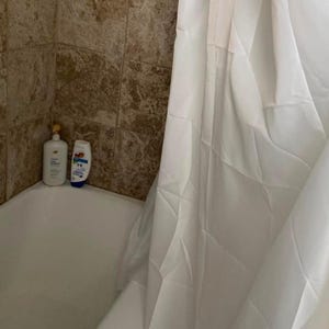 May include: A white shower curtain hangs in front of a white bathtub. Two bottles of bath products sit on the edge of the tub. The shower wall is tiled in shades of brown.