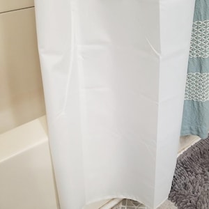 May include: A white shower curtain hangs in a bathroom, partially obscuring a light-colored tub. The curtain is made of a smooth, opaque material. The floor is tiled with a gray and white pattern, and a gray bath mat is visible.