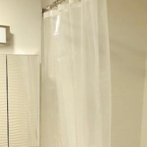 May include: A clear, transparent shower curtain hangs from a silver-colored rod. The curtain is made of a lightweight material and is slightly translucent, allowing light to pass through. The shower curtain is in a bathroom setting.