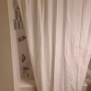 May include: A white shower curtain hangs from a metal rod with circular hooks. The curtain is partially drawn, revealing a white bathtub and shower fixtures. The bathroom walls are a light beige color.