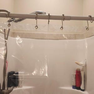 May include: A clear shower curtain hangs from a silver rod with metal hooks. The showerhead and faucet are visible. The shower curtain has a shelf with bottles of shampoo and other toiletries.