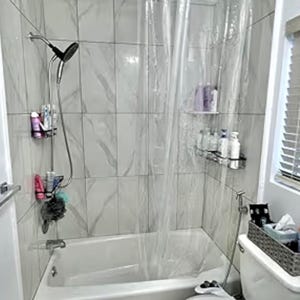 May include: A white bathtub and shower area with marble-patterned tiles. A clear shower curtain is drawn. A shower head and toiletries are visible. A toilet with a basket of supplies sits nearby.