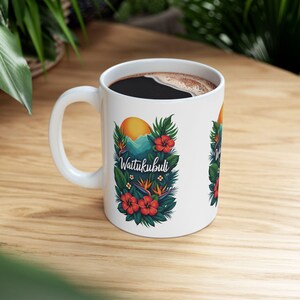 May include: White ceramic coffee mug with a handle, filled with dark liquid. The mug features a colorful design with the text "Waitukubuli", a sun, mountains, and tropical flowers in red, green, and orange.