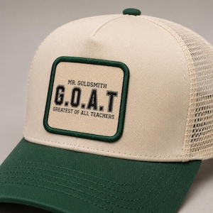 Puede incluir: Gorra de camionero beige y verde con parte trasera de malla. El parche frontal dice "MR. GOLDSMITH G.O.A.T GREATEST OF ALL TEACHERS" en texto negro. La gorra tiene una visera verde y un botón verde en la parte superior.