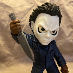 May include: A stylized vinyl figure of a character with a white mask, dark hair, and a navy blue jumpsuit. The figure has large, dark-rimmed eyes and is holding a gray plastic knife in a raised position, showcasing a detailed, sculpted appearance.