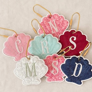 May include: A collection of seven shell-shaped fabric keychains in various colours including pink, teal, red, white, and navy blue. Each keychain features an embroidered letter in the centre and is attached to a gold-toned metal lobster clasp.