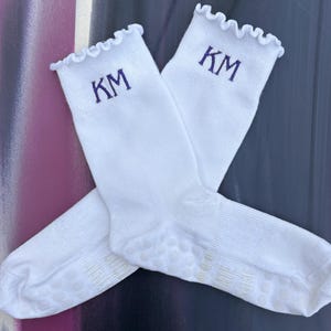 Op de afbeelding: Twee witte sokken met een gegolfde rand en de geborduurde letters "KM" in paars. De sokken zijn gekruist en hebben een grip patroon op de zolen. De achtergrond is een mix van paars, zwart en grijs.