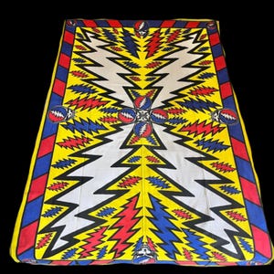 Puede incluir: Textil rectangular con un diseño vibrante de rayos y calaveras estilizadas. El patrón utiliza rojo, azul, amarillo y blanco, con un borde negro. El diseño es simétrico. Probablemente sea una manta o un tapiz.