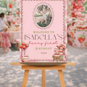 May include: A pink birthday sign on a wooden easel. The sign reads "Welcome to Isabella's berry first birthday 02.04.26" with a photo of a child. Strawberry and cake illustrations decorate the sign.