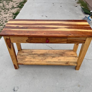 Puede incluir: Una consola de madera con acabado natural. La encimera presenta un patrón a rayas de tonos de madera claros y oscuros. Tiene un solo cajón con un pomo rojo y un estante inferior. La mesa está sostenida por patas anguladas.