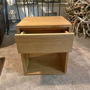 Puede incluir: Una mesita de noche de madera clara con un cajón parcialmente abierto. La mesita tiene un diseño simple y moderno con forma rectangular. El cajón es del mismo color que el resto de la mesita. La mesita está sobre una alfombra.