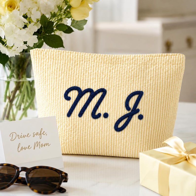 Puede incluir: Un estuche de seersucker amarillo y blanco con las iniciales M. J. bordadas en azul oscuro en la parte frontal. El estuche se muestra junto a una tarjeta blanca con el texto Drive safe, love Mom, unas gafas de sol y una caja de regalo.