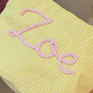 Puede incluir: Una tela de seersucker amarilla y blanca con el nombre Zoe escrito en letra cursiva. Las letras est&aacute;n formadas por un cord&oacute;n trenzado de color rosa claro que est&aacute; fijado sobre la superficie del material rayado y texturizado.