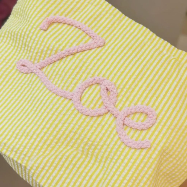 Puede incluir: Una tela de seersucker amarilla y blanca con el nombre Zoe escrito en letra cursiva. Las letras est&aacute;n formadas por un cord&oacute;n trenzado de color rosa claro que est&aacute; fijado sobre la superficie del material rayado y texturizado.