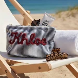 Puede incluir: Un estuche con cremallera de rayas azul marino y blancas con el nombre Khloe bordado en letra roja. El estuche contiene un tubo de protector solar SPF 50 y gafas de sol de carey, junto a una pinza para el pelo de leopardo.