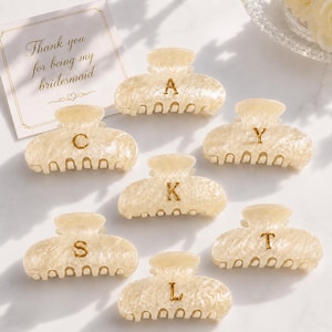 Puede incluir: Siete pinzas para el cabello de color crema y aspecto marmolado, cada una con una letra dorada: A, C, Y, K, S, T y L. Una tarjeta blanca con el texto 'Thank you for being my bridesmaid' se encuentra en la esquina superior izquierda.