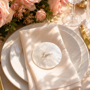 Könnte beinhalten: Ein kreisförmiges, weißes, schillerndes Platzkärtchen mit einer strukturierten, muschelartigen Oberfläche, auf dem der Name Lynn in eleganter goldener Schrift steht. Das Kärtchen liegt auf einer cremefarbenen Stoffserviette auf einem weißen Teller.