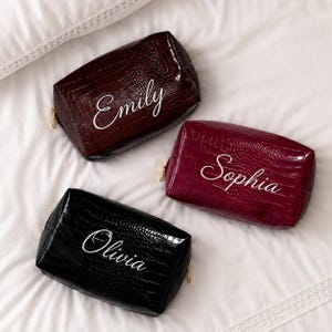 May include: Three rectangular cosmetic bags with a crocodile-embossed texture. The dark brown bag features the name Emily, the burgundy bag features the name Sophia, and the black bag features the name Olivia, all written in white script.