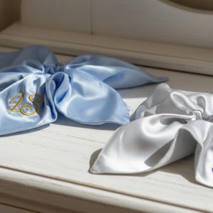 Może przedstawiać: Dwie satynowe kokardy do włosów w kolorze jasnoniebieskim i srebrnym. Jasnoniebieska kokarda ma złote haftowane cyfry "28". Kokardy ułożone są na białej powierzchni, z miękkimi fałdami i błyszczącą fakturą. Kokardy są prawdopodobnie na specjalną okazję.