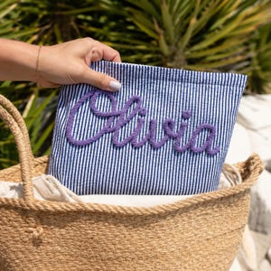 Könnte beinhalten: Eine rechteckige Tasche aus blau-weiß gestreiftem Seersucker-Stoff. Auf der Vorderseite ist der Name Olivia in erhabenen, violetten Buchstaben aus Kordelmaterial angebracht. Die Tasche befindet sich in einem geflochtenen Korb mit zwei Henkeln.