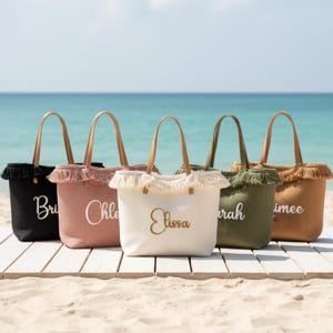Könnte beinhalten: Fünf personalisierte Tragetaschen in Schwarz, Rosa, Weiß, Grün und Beige, mit Namen in Weiß oder Gold gestickt. Jede Tasche hat eine Fransenborte und Ledergriffe. Die Taschen werden auf einem Holzsteg mit dem Meer im Hintergrund präsentiert.