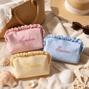 Puede incluir: Tres estuches rectangulares de tela con borde superior con volantes y estampado de cuadros vichy en rosa, azul y amarillo. Cada estuche tiene nombres bordados: Sophia en el rosa, Emma en el azul y Denise en el amarillo.
