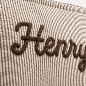 Puede incluir: Una superficie de tela acolchada con un patr&oacute;n de rayas verticales finas en marr&oacute;n y blanco. El nombre Henry est&aacute; bordado en el centro con letras cursivas gruesas de color marr&oacute;n oscuro, con aspecto de cuerda trenzada.