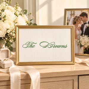 May include: A rectangular gold-toned frame containing a white fabric insert with the text "The Browns" embroidered in green cursive script. The frame features a beaded edge detail and is positioned on a light-colored wooden surface.
