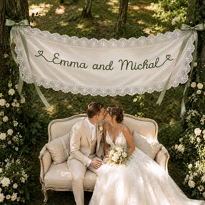 Może przedstawiać: Kremowy baner z tkaniny z napisem "Emma and Michal" ciemną czcionką, ozdobiony misternymi białymi koronkowymi brzegami i zawiązany pasującymi wstążkami na obu końcach, zawieszony między dwoma drzewami nad sofą w stylu vintage.