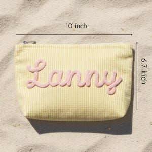 Puede incluir: Un estuche con cremallera de seersucker amarillo y blanco con el nombre Lanny en letras cursivas rosas en relieve. El estuche mide 25,4 cm de ancho y 17,0 cm de alto, con un tirador de cremallera de metal plateado en la esquina superior.