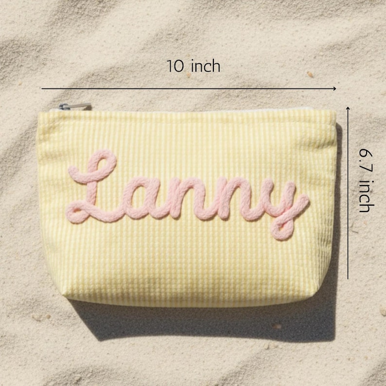 Puede incluir: Un estuche con cremallera de seersucker amarillo y blanco con el nombre Lanny en letras cursivas rosas en relieve. El estuche mide 25,4 cm de ancho y 17,0 cm de alto, con un tirador de cremallera de metal plateado en la esquina superior.