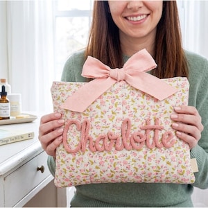 May include: A rectangular pillow with a floral pattern and the name "Charlotte" knitted in pink yarn. A large pink bow adorns the top. The background is a light, neutral tone.