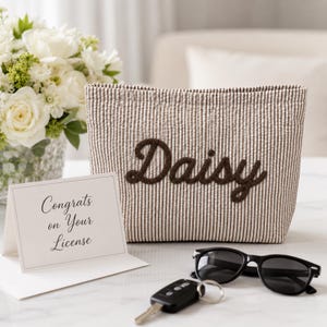 Puede incluir: Un estuche de tela a rayas beige y blancas con el nombre Daisy bordado en letra cursiva marr&oacute;n oscuro. Junto a &eacute;l hay una tarjeta blanca con el texto Congrats on Your License, gafas de sol negras y una llave de coche negra.