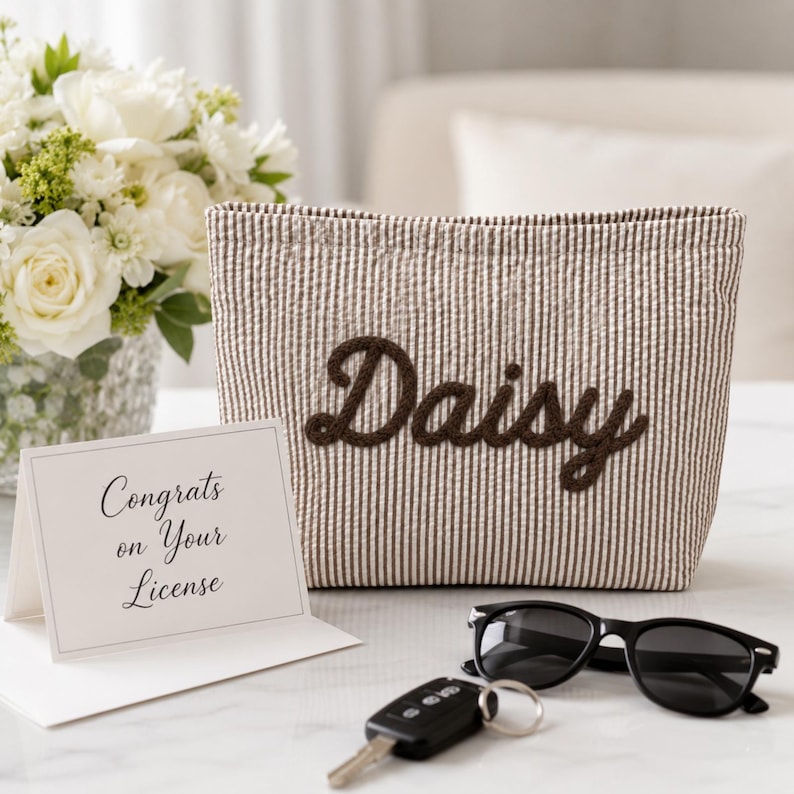 Puede incluir: Un estuche de tela a rayas beige y blancas con el nombre Daisy bordado en letra cursiva marr&oacute;n oscuro. Junto a &eacute;l hay una tarjeta blanca con el texto Congrats on Your License, gafas de sol negras y una llave de coche negra.