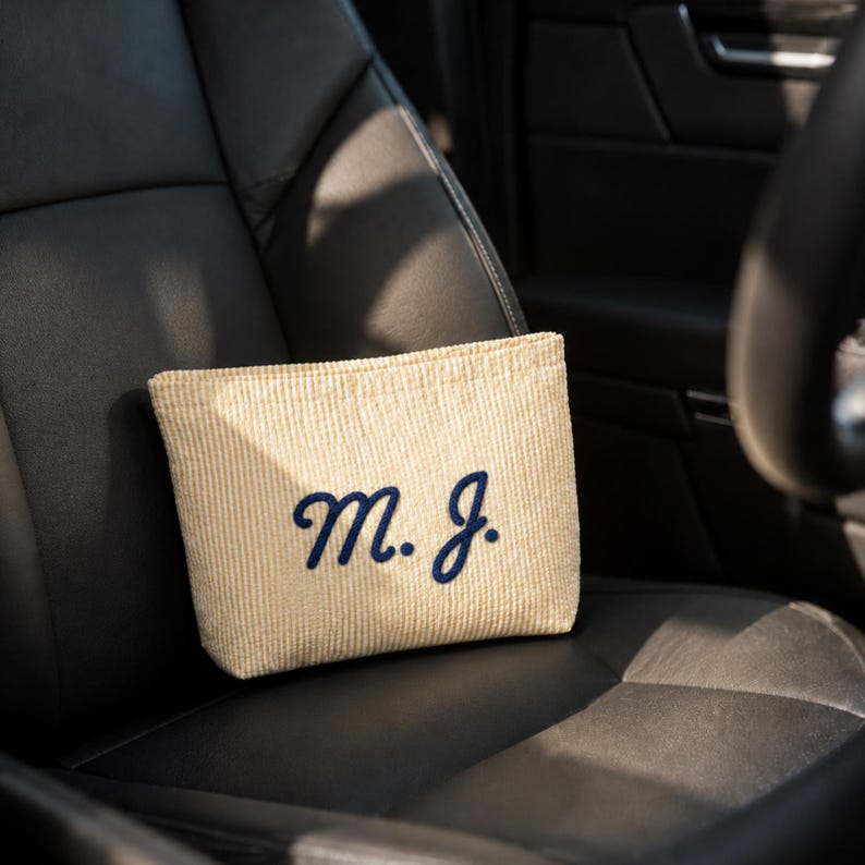 Puede incluir: Un estuche rectangular de rayas finas beige y blancas con las letras monograma M. J. bordadas en azul marino en una fuente cursiva. El estuche tiene una superficie de tela texturizada y est&aacute; colocado sobre un asiento de cuero negro.