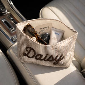 May include: A beige and white pinstriped fabric pouch featuring the name Daisy embroidered in dark brown script. The open pouch contains a pair of tortoiseshell sunglasses, a car key fob, a parking card, and a tube of Supergoop Glow Screen sunscreen.