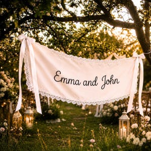May include: A cream-colored fabric banner featuring the black text Emma and John. The banner is edged with white lace trim and secured at both ends with long white satin ribbons tied to tree branches in an outdoor setting.