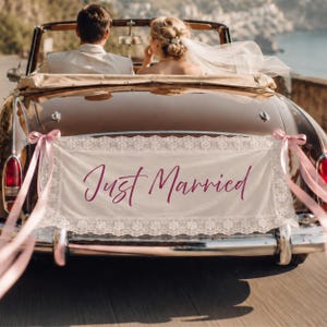 Może przedstawiać: Prostokątny baner z tkaniny w kolorze złamanej bieli z koronkowym wykończeniem i napisem Just Married ciemnor&oacute;żową kursywą, przymocowany do tyłu brązowego zabytkowego kabrioletu z jasnor&oacute;żowymi wstążkami po obu stronach zderzaka.