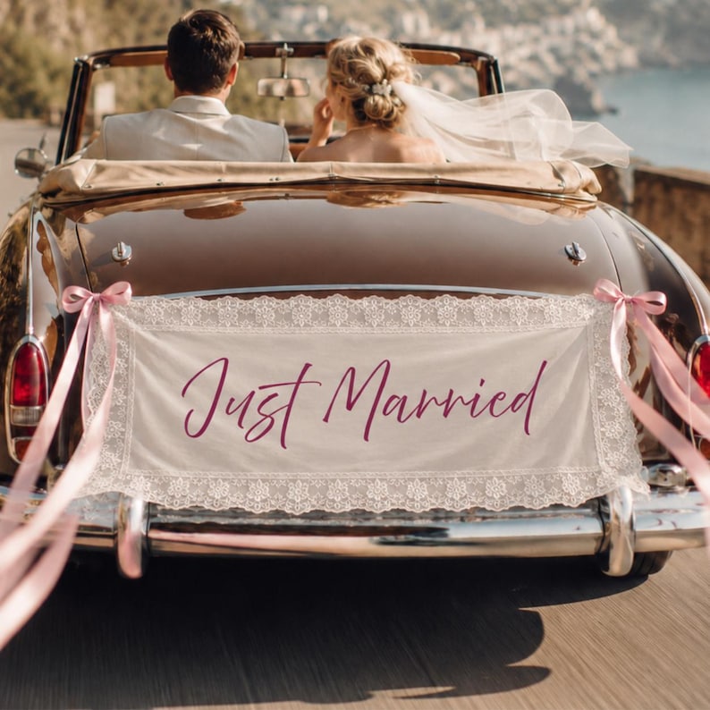 Może przedstawiać: Prostokątny baner z tkaniny w kolorze złamanej bieli z koronkowym wykończeniem i napisem Just Married ciemnor&oacute;żową kursywą, przymocowany do tyłu brązowego zabytkowego kabrioletu z jasnor&oacute;żowymi wstążkami po obu stronach zderzaka.