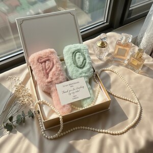 Può includere: Una confezione regalo contiene due articoli a forma di spazzola, soffici e personalizzati, uno rosa con una "P" lavorata a maglia e uno verde menta con una "O" lavorata a maglia. Una carta recita "TO MY BRIDESMAID, MIA, Thank you for standing by my side! love SARAH". Sono visibili anche una collana di perle e flaconi di profumo.