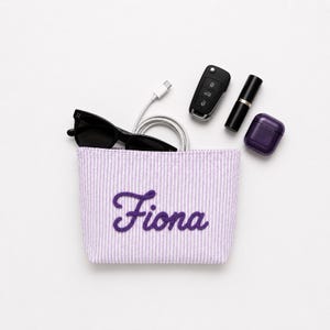 Puede incluir: Un estuche de tela a rayas de color violeta claro y blanco con el nombre Fiona bordado en letra cursiva de color violeta oscuro. El estuche contiene gafas de sol negras, un cable de carga blanco, una llave de coche, un pintalabios negro y un estuche de auriculares violeta.
