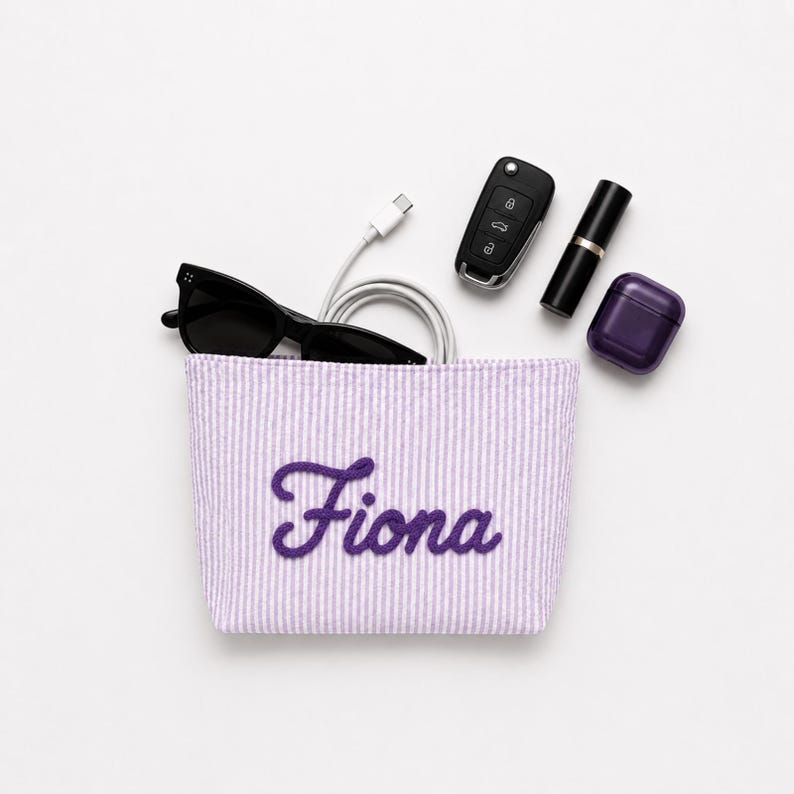 Puede incluir: Un estuche de tela a rayas de color violeta claro y blanco con el nombre Fiona bordado en letra cursiva de color violeta oscuro. El estuche contiene gafas de sol negras, un cable de carga blanco, una llave de coche, un pintalabios negro y un estuche de auriculares violeta.