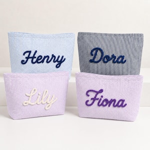 Puede incluir: Cuatro estuches rectangulares de tela con rayas verticales. El estuche superior izquierdo es azul claro con el nombre Henry en azul oscuro. El superior derecho es azul marino con el nombre Dora en marino. El inferior izquierdo es lavanda con el nombre Lily en blanco y el inferior derecho es lavanda con el nombre Fiona en morado.